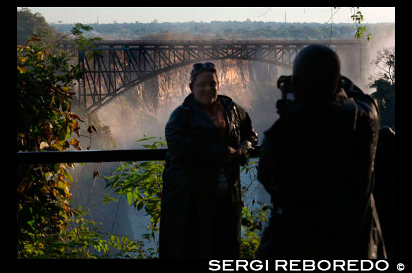 A couple takes photos in Victoria Falls. Behind the bridge between Zambia and Zimbabwe.  The Victoria Falls Bridge crosses the Zambezi River just below the Victoria Falls and is built over the Second Gorge of the falls. As the river is the border between Zimbabwe and Zambia, the bridge links the two countries and has border posts on the approaches to both ends, at the towns of Victoria Falls, Zimbabwe and Livingstone, Zambia. The bridge was the brainchild of Cecil Rhodes, part of his grand and unfulfilled Cape to Cairo railway scheme, even though he never visited the falls and died before construction of the bridge began. Rhodes is recorded as instructing the engineers to "build the bridge across the Zambezi where the trains, as they pass, will catch the spray of the Falls". It was designed by George Anthony Hobson of consultants Sir Douglas Fox and Partners, not as is often stated, Sir Ralph Freeman, the same engineer who contributed to the design of the Sydney Harbour Bridge. At the time of the design of the Victoria Falls Bridge, Freeman was an assistant in the firm who, in those pre-computer days, was calculating stresses. The bridge was constructed in England by the Cleveland Bridge & Engineering Company, before being shipped to the Mozambique port of Beira and then transported on the newly constructed railway to the Victoria Falls. It took just 14 months to construct and was completed in 1905. The bridge was officially opened by Professor George Darwin, son of Charles Darwin and President of the British Association (now the Royal Society) on 12 September 1905.[4] The American Society of Civil Engineers lists the bridge as a Historic Civil Engineering Landmark. The Victoria Falls Bridge did not bring the first train or the first railway to Zambia. To push on with construction of the railway north as fast as possible, Cecil Rhodes insisted that the Livingstone to Kalomo line be laid before the bridge was finished. Then a locomotive was conveyed in pieces across the gorge by the temporary electronic cableway used for the transportation of the bridge materials and nicknamed the 'Blondin' by the construction engineers. The locomotive was re-assembled and entered service months before the bridge was complete.