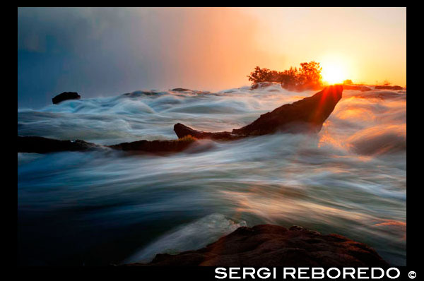 Posta de sol a les Cascades Victòria. Les cascades Victòria o el Mosi-oa-Tunya (Tokaleya Tonga: el fum que trona, la 'i' és silenciosa) és una cascada situada al sud d'Àfrica en el riu Zambezi entre els països de Zàmbia i Zimbabwe. Les caigudes es creu que són els més grans del món en termes de volum d'aigua que passa a través. Aquests antiga i realment espectacular Falls es van donar a conèixer a la resta del món en 1855 per l'explorador escocès extraordinari, David Livingstone. Livingstone havia estat tractant de trobar una ruta a la costa est del continent africà. Viatjant cap al sud a l'est de Luanda a Sesheke, es va trobar amb aquesta, la més magnífica cascada i va canviar el nom a les Cascades Victòria després que el monarca britànic, la reina Victòria. Livingstone es va dur a les cataractes per les tribus Makalolo persones en una piragua. Durant el Període Juràssic (fa 150-200 milions d'anys), l'activitat volcànica va donar lloc a dipòsits de basalt gruixudes que cobreixen grans parts del sud d'Àfrica. A mesura que la renta es refreda i es solidifica, van aparèixer esquerdes a l'escorça basàltica dur, que s'omple de fang i calç. L'erosió i el curs del poderós riu Zambezi tallen a través d'aquests materials més tous, formant la primera d'una sèrie de cascades. Durant almenys 2000 anys, les caigudes han retrocedit 8 quilòmetres aigües amunt, com el Zambezi tallar el seu camí a través de set goles. Aquesta història geològica es pot veure al basalt fosc en la sèrie de goles rocoses sota les cataractes. Es creu que la Cataracta del Diable, que és actualment el punt més baix de les Cataractes Victòria, amb el temps es convertirà en el pròxim barranc com el riu continua tallant el seu camí de retorn riu amunt. En essència, el riu cau en un barranc just al davant de les cataractes, i després flueix a través d'un tall estret. Vostè pot veure les cascades recte des de l'altre costat del barranc.