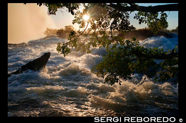 Posta de sol a les Cascades Victòria. Victoria Falls és el resultat de la pedra arenisca suau que omple enormes esquerdes a la dura roca basàltica de l'altiplà. A mesura que el Zambezi superior fluir a través de l'altiplà en l'antiguitat, es van trobar les esquerdes i va començar a portar lluny la roca més suau, creant finalment una sèrie de goles. Els geòlegs estimen que el riu ha anat disminuint en aquests barrancs durant almenys els últims 100.000 anys. A mesura que la roca es desgasta, la cataracta segueix aquestes esquerdes gegantines i es mou riu amunt a través de l'altiplà. Dr David Livingstone havia estat viatjant pel riu Zambezi al sud d'Àfrica des de fa diversos mesos. Potser un centenar de quilòmetres riu amunt durant aquesta expedició havia arribat a Ngonye Falls. Livingstone havia trobat la cataracta és molt impressionant. Les cataractes eren només uns 60 metres d'altura sinó que es va estendre a tot el llarg del riu ample i la quantitat d'aigua que es va precipitar sobre ells va ser sorprenent. No obstant això, en aquest dia, 17 novembre de 1855, s'ha dit que seria veure una cascada espectacular que deixarien petit el de Ngonye. Livingstone, igual que la majoria dels europeus, tenia alguns dubtes sobre això. Després de tot, en aquest punt el riu fluïa mandrosament a través d'un altiplà plana sense una muntanya o una vall a la vista. Com pots tenir una gran cascada i sense una característica de la terra que podria causar un sobtat canvi en l'altura del riu? Com la canoa estaven remant en aigües avall de cap, però, Livingstone va començar a sentir una remor llunyà. Per davant un núvol de boira va sorgir d'aparentment enlloc. En el moment de la canoa es va aturar en una petita illa i Livingstone havia caminat fins a la vora de la cataracta, el so es va convertir en un rugit ensordidor.