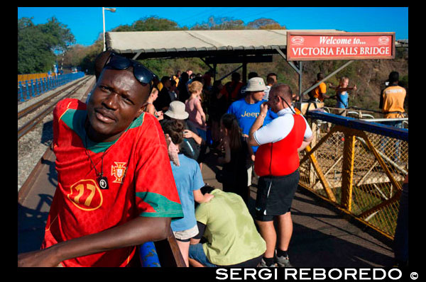 La gente alrededor de Victoria Falls Bridge. The Victoria Falls Puente cruza el río Zambezi justo debajo de las Cataratas Victoria y está construido sobre la Segunda Quebrada de las caídas. A medida que el río es la frontera entre Zimbabwe y Zambia, el puente une los dos países y cuenta con puestos fronterizos en los accesos a los dos extremos, en las ciudades de Victoria Falls, Zimbabwe y Livingstone, Zambia. Durante más de 50 años, el puente fue cruzado con regularidad por los trenes de pasajeros como parte de la ruta principal entre la entonces Rhodesia del Norte, África del Sur y Europa. Los trenes de carga realizan principalmente mineral de cobre (más tarde, lingotes de cobre) y madera de Zambia, y el carbón en el país. La edad de los problemas de puentes y mantenimiento han llevado a restricciones de tráfico en horas. Los trenes se cruzan en menos de caminar ritmo y camiones fueron limitados a 30 t, necesitando camiones más pesados ??que hacer un largo desvío a través de la Kazungula Ferry o puente Chirundu. El límite se elevó después de las reparaciones en 2006, pero más fundamental la rehabilitación o construcción de un nuevo puente ha sido ventilado. Durante la crisis de Rhodesian UDI y la guerra de Bush, el puente fue cerrado con frecuencia (y los servicios regulares de pasajeros no se han reanudado con éxito). En 1975, el puente fue el escenario de las conversaciones de paz fallidos cuando las partes se reunieron en un vagón de tren suspendido sobre el barranco de nueve horas y media. En 1980 los servicios de transporte de mercancías por carretera y se reanudaron y han continuado sin interrupción, excepto para el mantenimiento.