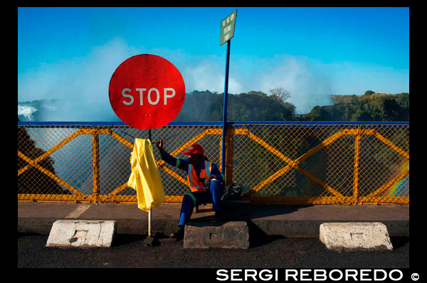 Pare la muestra en el puente fronterizo Zambia y Zimbabwe. El Puente de Victoria Falls es de 100 años de edad en 2005. El Puente de Victoria Falls fue la creación de Cecil Rhodes, una característica clave en su sueño de un Cabo de ferrocarril de El Cairo, a pesar de que él nunca visitó las Cataratas Victoria y murió dos años antes de que el ferrocarril llegó a ellos - antes había comenzado la construcción del puente. El levantamiento preliminar de las bases para el puente se hizo en 1900-1901, durante la época de la Guerra Boer estaba en su apogeo, hacia el sur de comunicaciones fueron cortadas, y la construcción de la vía férrea a Victoria Falls fue mucho retraso, pero nunca lo suspendieron, a lo largo militar operaciones. La arena de la guerra no incluyó Rhodesia, y la obra de la construcción del ferrocarril nunca cesó durante todo el período. En 1900 se le pidió Rodas escribir un delantero para el libro 'De Cabo a El Cairo "por Grogan y Sharp. Ewart de Scott Grogan, junto con Harry de Sharp emprendió el viaje por tierra épica desde el Cabo hasta El Cairo, aunque Grogan fue el único en completar todo el viaje, y así convertirse en el primer hombre en alcanzar tal empresa. Ellos viajaron en tren, barco y otros medios donde podían, pero caminaron durante gran parte de su viaje a través del continente africano. Inspirado por la lectura de Frederic Courtney Selous de 'Andanzas de un cazador en África', Grogan se propuso demostrar su valía y ganar la mano de su amor en el matrimonio. Su viaje duró tres años, alcanzando Grogan Cairo en 1900. Sir Charles Metcalfe, un amigo personal cercano de Rodas, siguió a sus deseos y decidido a buscar el puente justo por debajo de las cataratas. Él llevó a cabo los exámenes preliminares del sitio en junio 1901, antes de regresar a Gran Bretaña para recaudar fondos para el proyecto. En 2005 se llevó a cabo una importante encuesta 100 años del puente. Informes previos de funcionarios de los Ferrocarriles Nacionales de Zimbabwe (NRZ) habían planteado el peligro que representa el puente de carga pesada y estaba cerrado al tráfico pesado por más de un año día para permitir el trabajo de emergencia. Un informe, publicado en enero de 2005 por NRZ destacó "vibraciones excesivas hacen sentir cada vez que un camión pesado atraviesa el puente". Durante los trenes restricciones cruzadas a menos que el ritmo de caminar y camiones estaban limitados a una carga de 30 toneladas, lo que exige camiones más pesados ??que hacer un largo desvío a través de la Kazungula Ferry o puente Chirundu. La elección para los ingenieros era o bien para reconstruir o reforzar el puente, pero se conformó con el refuerzo. Después de las reparaciones que cuestan EE.UU. $ 1.7 millones el puente fue reabierto al tráfico pesado el 15 de junio de 2006, y ahora puede soportar cargas de hasta 56 toneladas para los próximos cinco años. Durante este período, más reparaciones se llevarán a cabo para que el puente para sobrevivir otro siglo. Reemplazo del puente con una moderna estructura similar se ha estimado que costará más de 32 millones de dólares EE.UU.. En noviembre de 2010 se anunció que un peaje en el Puente de Victoria Falls se estaba estudiando en un intento de recaudar los fondos necesarios para el mantenimiento. Las recomendaciones formuladas por los consultores internacionales han dicho que podría durar otros 100 años si se mantienen adecuadamente. Se ha estimado que más de 1,9 millones de dólares EE.UU. se necesita para inyectar en el mantenimiento del puente - EE.UU. $ 800,000 serán para la actualización de la plataforma ferroviaria; 300.000 dólares EE.UU. para la actualización de la plataforma de acera y 800.000 dólares EE.UU. para la actualización de la cubierta de carretera. Poseído originalmente por Rhodesia Ferrocarriles, Victoria Falls Bridge es ahora propiedad conjunta de los ferrocarriles nacionales de Zambia y Zimbabwe. Está gestionado por la Ferrocarriles Surgió Properties (Private) Limited (ERP), una compañía interestatal de propiedad conjunta de los gobiernos de Zimbabwe y Zambia. No hay servicios regulares de viajeros por ferrocarril sobre el puente en la actualidad. Sin embargo, el vapor arrastrado excursiones en un histórico salón comedor se ofrecen diariamente entre la estación de Victoria Falls (Zimbabwe) y Livingstone (Zambia). Además, varios trenes de cruceros de lujo hacen el viaje desde África del Sur en cuanto a Livingstone, cruzando el puente como parte de su viaje. Hoy en día el Victoria Falls Bridge es la ubicación para el salto bungee Shearwater 111 metros, que inició operaciones en 1993. El Jefe Mukuni locales apareció en el puente en traje de gala, acompañado por la mayoría del pueblo, a tomar su turno para saltar. Después de varios intentos fallidos que derrocó al vacío, con el acompañamiento de vítores de los espectadores. Se ha afirmado que esta pies le deben pagar un lugar en el Libro Guinness de los Récords como el primer jefe africano que bungeed en la historia de este deporte. Más de 50.000 personas se han comprometido a la emoción de saltar desde el puente sin ningún incidente. La operación se ha expandido para incluir un columpio en bungee y tirolesa. En 2010 una plataforma de la visión renovada, restaurante y bar, así como un museo interpretativo, se abrió en el lado de Zambia. La última actividad turística en el puente son excursiones de puentes históricos interactivos, donde los grupos están guiados bajo el puente utilizando arneses de seguridad y cuerdas.