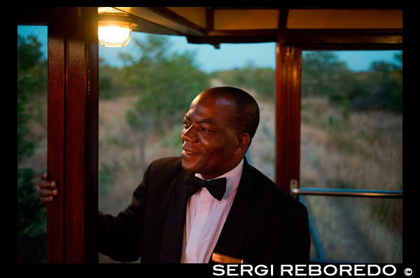 Uno de los trabajadores ferroviarios del tren de lujo Livingstone Express. Cena de lujo se redefine cuando el lugar es el vagón comedor de un tren a vapor de 1920, en medio de la sabana de Zambia. El Royal Livingstone Express es una empresa conjunta entre Bushtracks África y Sun International. Esta experiencia única es todo incluido y los huéspedes son trasladados de todo Livingstone y las casas de campo de los alrededores de las antiguas oficinas de la línea Mulobezi en la ciudad de Livingstone. Subiendo al tren en una alfombra roja le permitirá disfrutar de esta experiencia que hay que hacer. Cada huésped es recibido personalmente y ofreció una bebida de bienvenida al subir a bordo del carro de la vendimia inmaculada Salón de primera clase. El Royal Livingstone expreso consta de cinco vagones, restaurado por Rohan Vos de Rovos Rail y éstos incluyen dos coches comedor, un coche del club / cocina, alquiler de sala de estar, y un coche de observación y se tira ya sea por una clase 10 N º 156 o un 12 Clase N º 204 locomotoras. Una vez que todos los clientes están a bordo del viaje comienza a través suburbio Dambwa hacia el Parque Nacional de Mosi-oa-Tunya, que corre paralela al río Zambezi. Las bebidas se sirven mientras que usted hace el juego viendo desde la comodidad de su asiento en uno de los salones o en el coche de observación. Una vez que el sol se ha puesto espectacularmente, el gerente de tren invita a los huéspedes a cenar. Los coches comedor están revestidos de brillantes paneles de madera y luz de la lámpara suave realza la elegancia de los cubiertos de plata, cristalería y ropa de damasco. Una vez sentado, curso tras curso delicioso aparecen como por arte de magia. El servicio en el tren es impecable y la comida simplemente divina. (Asegúrese de que venga con hambre!) Durante la cena de seis platos del tren empuja de nuevo hacia Livingstone y muy pronto llega de vuelta en el Mulobezi Siding aproximadamente a las 21h30, donde los huéspedes felices desembarcan y son transferidos de vuelta a sus hoteles.