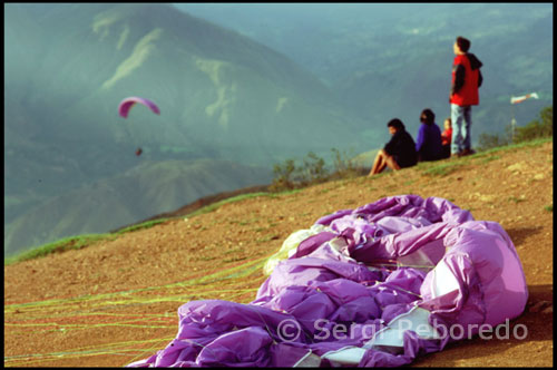 Algunos aficionados se acercan para ver volar los parapentes.  Parapente en Tierra Negra.  Venezuela. Esa misma tarde salgo del centro de Mérida después de comer. La idea es hacer parapente en una montaña cercana. Después de 45 minutos de tortuoso recorrido, por fin habíamos llegado a Las González o al lugar más comúnmente llamado Tierra Negra. Nadie supo explicarme el porqué de este nombre, pero la cuestión es que la tierra no era ni mucho menos de color negra, sino de un tono rojo cobrizo que contrastaba enormemente con el azul electrizante del cielo. Nos encontrábamos en una enorme planicie a 1700 metros sobre el nivel del mar con unas fabulosas vistas del valle y del río Chama.   El rango máximo y mínimo de velocidades relativas para un modelo de parapente intermedio es: Velocidad máxima: 50 kilómetros/hora. Velocidad mínima: 24 km/h, un parapente de competición puede superar los 60 kilómetros/hora y una relación de planeo, o "fineza" de 10:1, es decir, que por cada 10 metros relativos de aire que avanza se desciende uno (debemos pensar que estando en el aíre, la velocidad en este caso siempre será relativa a la fuerza y dirección del viento, y no confundir con la velocidad real a la tierra GPS). Se trata de poder despegar, volar y aterrizar con un ala flexible por los propios medios del piloto, es decir; a pie. El ideal sería desde una ladera no muy inclinada y encarada a un viento moderado de unos 10-20 km/h, o menos aún si se pretende volar cerca de la costa, ya que el aire tendrá una mayor densidad allí debido a unas presiones atmosféricas y humedades relativas más elevadas. Después el piloto se sentará cómodamente en una silla o arnés, unidos a las bandas de suspentaje mediante dos mosquetones.