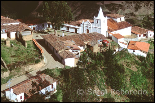 Iglesia y alrededores. Los Nevados.  Venezuela. Varios excursionistas que vuelven de regreso se cruzan en mi camino. Van provistos de caballos o mulas, ya que, tanto a pie del funicular como en el pueblo, es posible alquilarlos si no tenemos la forma física adecuada para esta caminata de cinco horas. El paisaje montañoso me cautiva por su abrumadora belleza, en la que destaca sobre todo el contraste del color cobrizo del páramo con amarillo de los frailejones. Esta planta típica andina, de flor parecida a la margarita, es de la familia de las compuestas y florece hacia el mes de Septiembre inundando el páramo de un color dorado intenso. El camino es largo y atraviesa varios riachuelos que transportan las gélidas aguas de las montañas cercanas hacia las planicies. Los Nevados en Mérida es un pequeño pueblo edificado sobre las estribaciones que descienden del pico El Toro, en la margen derecha del río de Nuestra Señora. Está ubicado a una altura de 2.400 msnm, frente a una sucesión de montañas que se extiende en la lejanía hasta los llanos de Barinas. Su temperatura media anual es de 15º C. Su población no sobrepasa los 150 habitantes. Los antiguos pobladores de Los Nevados eran indígenas que dejaron fiel testimonio de la antigua civilización mirripuy en los andenes o terrazas de siembra, así como en el camino empedrado que conduce hasta el pueblo y en las técnicas de construcción todavía vigentes en el lugar.